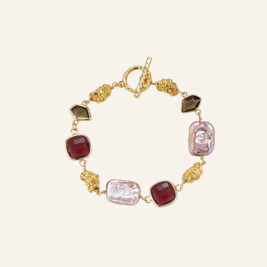 18k gold-plated copper bracelet with burgundy stones, smoky quartz, baroque pearls and ornate golden charms on white background.