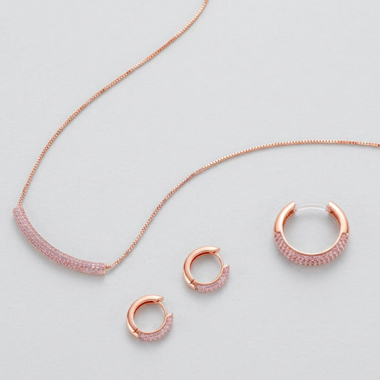 Rose gold pavé zirconia bar necklace, hoop earrings, and open ring set with pink stones on cream background.