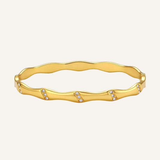 Gold-toned wavy bangle with bamboo-style segments and CZ crystal clusters on cream background. 18K plated stainless steel.