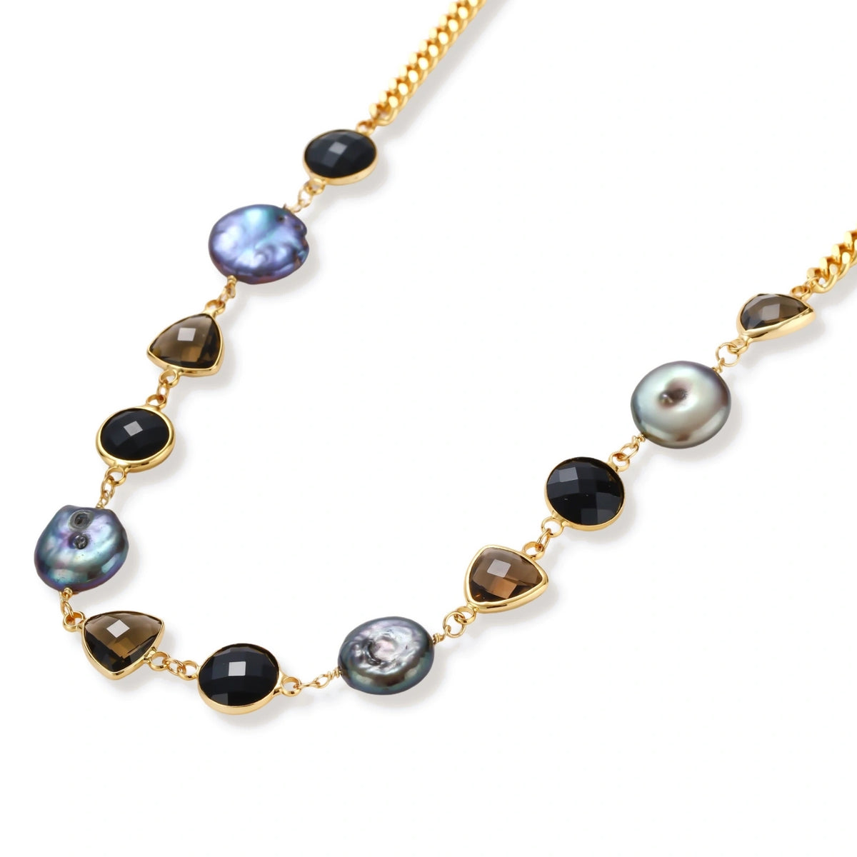 Gold plated curb chain necklace with iridescent baroque pearls, black round stones and smoky triangular stones on a cream background.