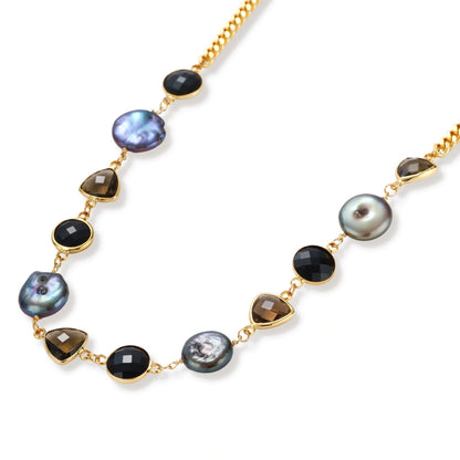 Gold plated curb chain necklace with iridescent baroque pearls, black round stones and smoky triangular stones on a cream background.